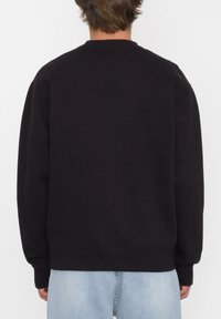 Sudadera negra con cuello redondo, puños y dobladillo de canalé. Hecha de una tela texturizada, vista desde atrás, combinada con jeans de mezclilla claros.