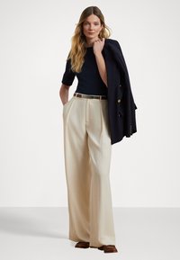 Pantalons larges taille haute crème avec des plis, associés à un haut ajusté bleu marine et une veste bleu marine drapée sur une épaule. Chaussures brunes.