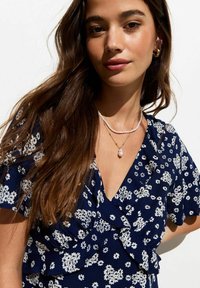 Vestido floral de marina con patrón blanco, escote en V, detalles con volantes, combinado con collares en capas. Textura de tela suave.