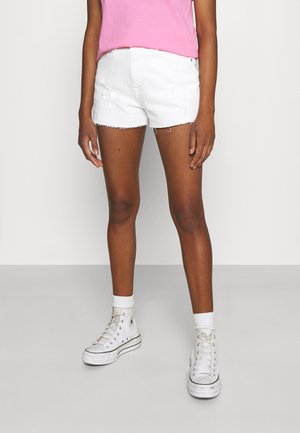 Person iført hvide denimshorts, hvide høje sneakers, hvide sokker og en pink top, stående på en grå gulv mod en hvid væg.