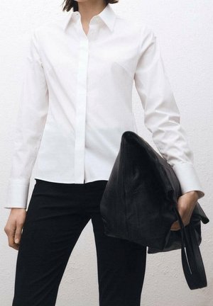 Nainen, joka pukeutunut istuvaan valkoiseen napitettuun paitaan ja mustiin housuihin, pitää oikeassa kädessään suurta mustaa nahkalaukkua.