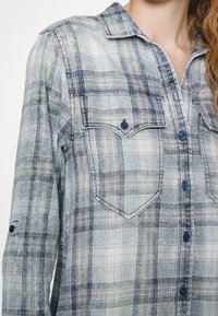 Jasnoniebieska koszula w kratę z niebieskimi guzikami, długimi rękawami i dwoma kieszeniami na piersi. Wykonana z materiału przypominającego dżins, o miękkiej teksturze.
