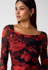 Zwarte mesh top met een rood bloemenpatroon, voorzien van lange mouwen en een vierkante halslijn. Een gouden ketting voegt een contrasterend detail toe.