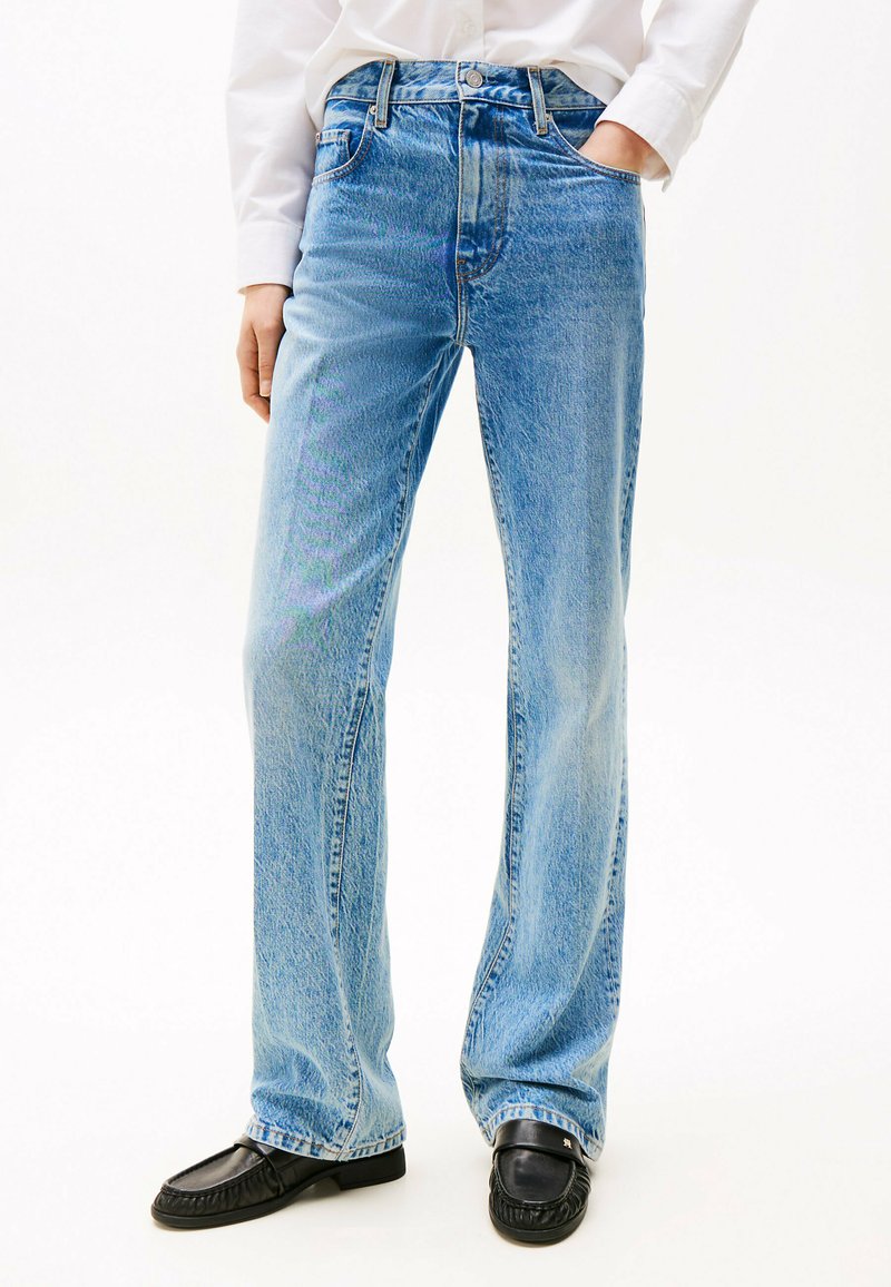 Jeans en denim bleu clair avec une taille haute, une coupe large et une finition délavée, comportant cinq poches et une fermeture à bouton classique. Associés à des mocassins noirs.