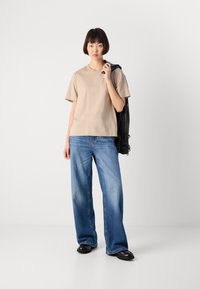 Giovane donna in piedi con una maglietta beige a maniche corte e jeans a gamba larga blu, che tiene una giacca di pelle nera appoggiata sulla spalla.