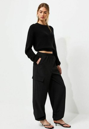 Donna che indossa un maglione nero corto, pantaloni cargo neri larghi e tacchi neri open toe, in piedi su uno sfondo bianco semplice.