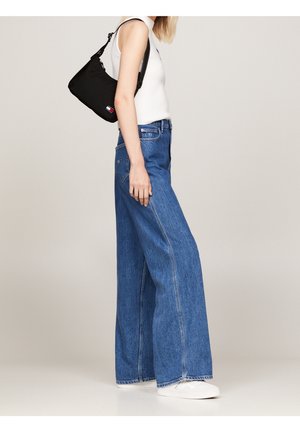 Jean large bleu en denim, associé à un haut sans manches blanc et un petit sac à épaule noir avec un logo. Chaussures blanches visibles.