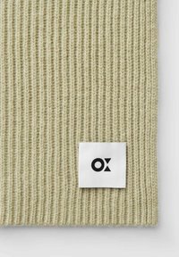 Gebreid accessoire in lichtgroen met ribbelstructuur, met een wit vierkant label met een zwart logo.
