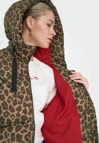Padded jacket in a brown leopard print, featuring a hood and black zipper. The interior is red. A white shirt with red print is visible underneath.