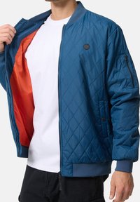 Man met blauwe doorgestikte bomberjas met oranje voering, wit T-shirt en zwarte broek, die de jas openhoudt om de binnenvoering te tonen.