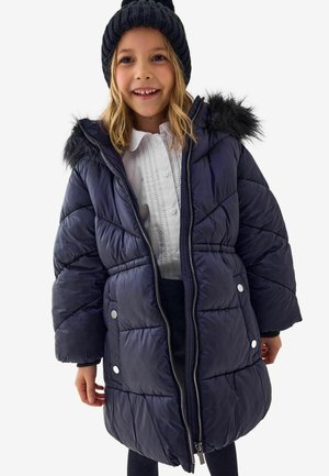 Veste matelassée navy avec une fourrure synthétique sur la capuche, fermeture éclair et détails à boutons pression. Portée sur une chemise blanche.
