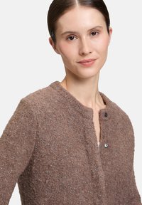 Frau mit braunem, zurückgebundenem Haar, die eine strukturierte braune Strickjacke mit silbernen Fadenakzenten und Druckknöpfen trägt, frontal blickend mit einem leichten Lächeln.