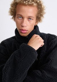 Pull en polaire noir avec un col haut, tissu texturé et une demi-fermeture éclair. Présente une coupe décontractée avec des poignets élastiques.