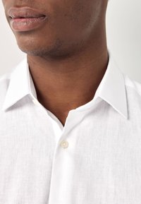 Persona dalla pelle scura che indossa una camicia di lino bianca con il primo bottone sbottonato, mostrando parte del collo e della parte inferiore del viso.