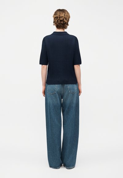 Άτομο με κοντά σγουρά μαλλιά στέκεται με την πλάτη προς τα εμπρός, φοράει μπλε κοντομάνικο πουκάμισο και φαρδιά μπλε τζιν, πάνω σε λευκό φόντο.