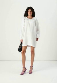 Vestido branco de linho com mangas longas e bufantes e decote redondo. A modelo segura uma mala preta e usa sandálias de salto alto roxas.