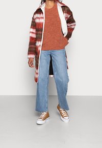 Veste extérieure à carreaux rouge, blanche et verte avec un intérieur duveteux, associée à un pull rouille, un jean large bleu et des baskets marron.