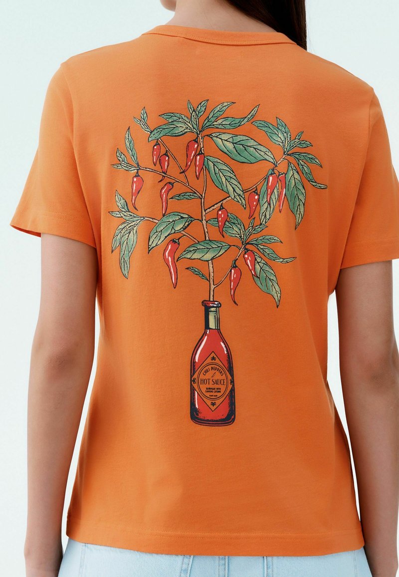 Oranssi puuvillapaita, jossa on graafinen kuvio kuumakastelaposta ja chilipensasta vihreänä ja punaisena takana.