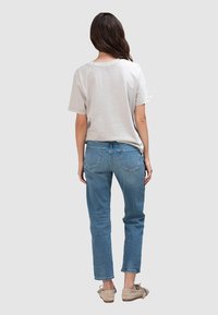 Lichtgrijs, katoenen T-shirt met korte mouwen, gecombineerd met blauwe spijkerbroek met achterzakken en een ontspannen pasvorm. Draagt beige loafers.