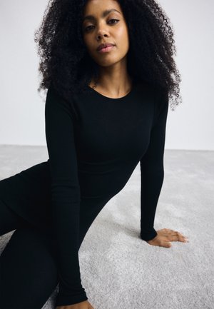 Svart langermet bodysuit med rund hals. Modellen har krøllete hår og sitter på en lys teksturert overflate, med en nøytral bakgrunn.