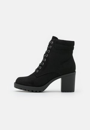 Bottine noire en daim avec talon haut épais, laçage devant avec œillets métalliques et semelle à crampons robustes, vue de profil latéral.