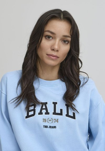 Lys blå sweatshirt med rund hals, med svart og gull tekst "BALL 1974 THE JEANS." Myk stoff med en avslappet passform.
