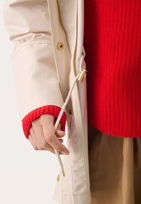 Beige välimantel kuldsete detailidega, millel on reguleeritav nöör ja punased kudumist käeaugud. All on tekstuuriga punane sviiter.