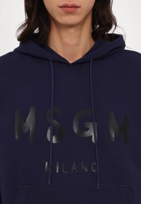 Personne portant un sweat à capuche bleu marine avec des inscriptions noires brillantes "MSGM" et "Milano" sur le devant, ainsi que des cordons de capuche qui pendent.