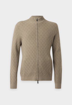 Mann trägt einen beigen, diamantgemusterten Reißverschluss-Pullover, schwarzes Hemd, lockere schwarze Hose und schwarze Schuhe, steht mit einer Hand in der Tasche.