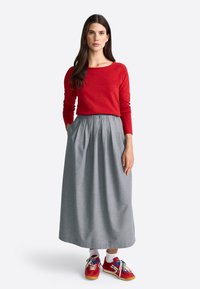 Vrouw in een rode langmouwen top, grijze maxi rok en rode sneakers met witte sokken, staande met haar handen in de zakken van de rok.