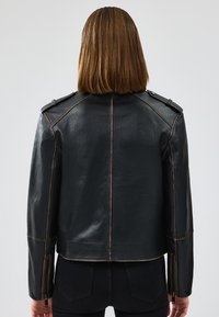 Giacca in pelle nera con spalline, cuciture visibili e polsini con zip. La vista posteriore mostra un design aderente e una texture liscia.