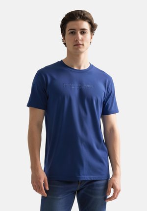 Jeune homme debout, de face, portant un t-shirt bleu Armani Exchange et un jean bleu foncé sur un fond clair uni.