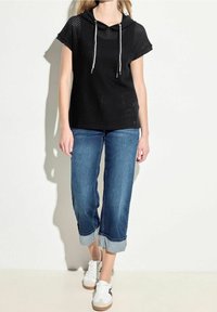 Czarna perforowana bluzka z kapturem i krótkimi rękawami, zestawiona z dżinsami w niebieskim kolorze z podwiniętymi mankietami oraz białymi sneakersami. Tekstura dodaje głębi całemu zestawowi.