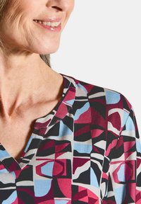 Bunt gemusterte Bluse mit geometrischem Muster in Rot, Blau, Grau und Schwarz. V-Ausschnitt-Design mit glatter Stoffoberfläche und sichtbaren Nähten.