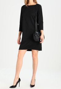Robe longue noire à manches longues avec un décolleté bateau, longueur genou, en tissu doux ; associée à des talons hauts noirs et à un petit sac en bandoulière.