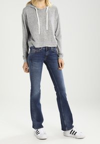 Sudadera gris con cordones blancos, combinada con jeans de campana azules y zapatillas blancas con rayas negras. Atuendo simple y casual.