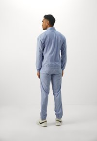 Mand, iført en lys blå Lacoste træningsdragt og hvide Nike sneakers, står med ryggen til en enkel hvid baggrund.