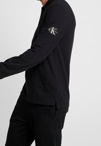 Černá polokošile s dlouhým rukávem, dvoupohybovým zapínáním, bočními rozparky a nášivkou loga Calvin Klein Jeans na levém rukávu. Bavlněná látka.