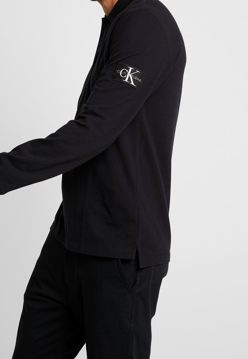 Černá polokošile s dlouhým rukávem, dvoupohybovým zapínáním, bočními rozparky a nášivkou loga Calvin Klein Jeans na levém rukávu. Bavlněná látka.