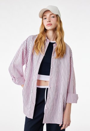 Młoda osoba w białej czapce, czarnym krótkim topie, luźnej różowo-białej koszuli w paski na guziki i ciemnych spodniach na białym tle.