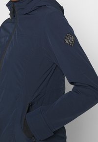 Námořnická modrá bunda se zipovým uzávěrem, dlouhými rukávy, texturovanou tkaninou a trojúhelníkovou černou nášivkou na horní části rukávu.