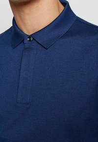 Mörkblå pikétröja med en texturerad tyg, med klassisk krage, tvåknappling och kontrasterande knapp för detalj.