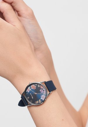 Navy silicone watch features a round, silver case with a floral design on a dark face; numerals at 12, 3, 6, and 9 positions.