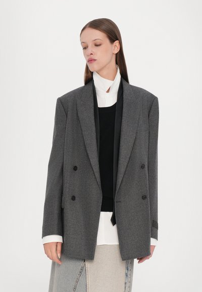 Blazer grigio doppiopetto realizzato in tessuto texturizzato, con revers a punta e bottoni neri, sovrapposto a una maglietta nera e una camicia bianca.