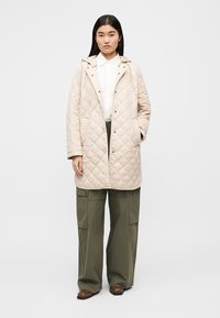 Ανοιχτό μπεζ παλτό με κουκούλα, κουμπιά στο μπροστινό μέρος με πατιλέτα και δύο πλαϊνές τσέπες. Συνδυασμένο με χακί παντελόνι καμπάνα τύπου cargo και καφέ παπούτσια.