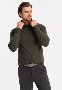Un pull vert foncé avec un décolleté décontracté, des manches ajustées et des poignets côtelés, associé à un pantalon gris et une ceinture brun clair.