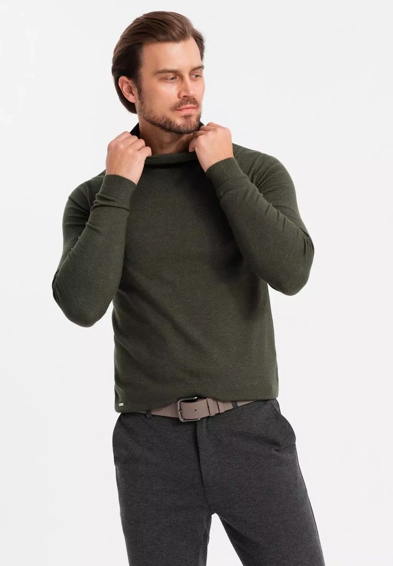 Un pull vert foncé avec un décolleté décontracté, des manches ajustées et des poignets côtelés, associé à un pantalon gris et une ceinture brun clair.