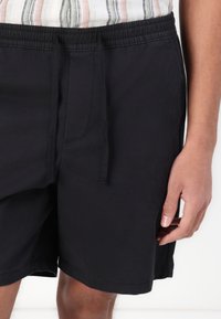 Sorte elastiske shorts med snøring i livet, med sidelommer og en myk tekstur. Oppbrett ved kanten, uten mønstre.