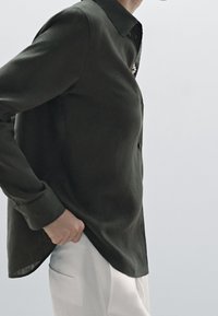 Chemise boutonnière à manches longues de couleur vert foncé en tissu léger, avec un col pointu et une texture subtile. Associée à un pantalon blanc.