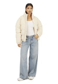 Puchowa kremowa kurtka ze sztruksu z wysokim kołnierzem, zestawiona z jasnoniebieskimi szerokimi spodniami typu jeans. Modelka ma na sobie biały top i białe trampki.